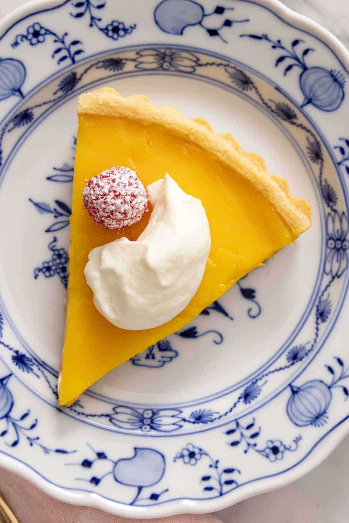 Overhead shot of a slice of lemon tart, topped with a berry and whipped cream, on a blue and white plate.