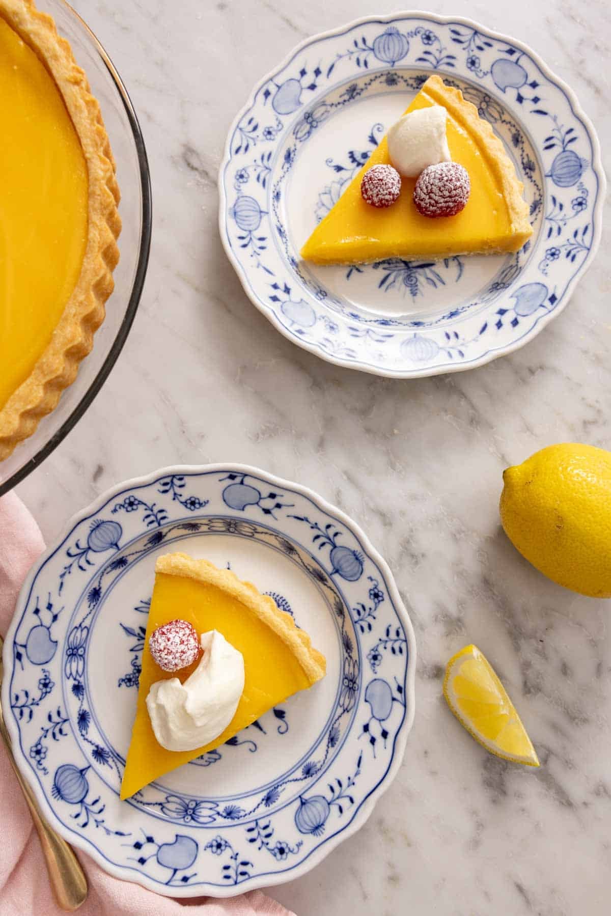 Overhead shot of two slices of lemon tart on a marble countertop with a whole lemon and slice of lemon next to them.