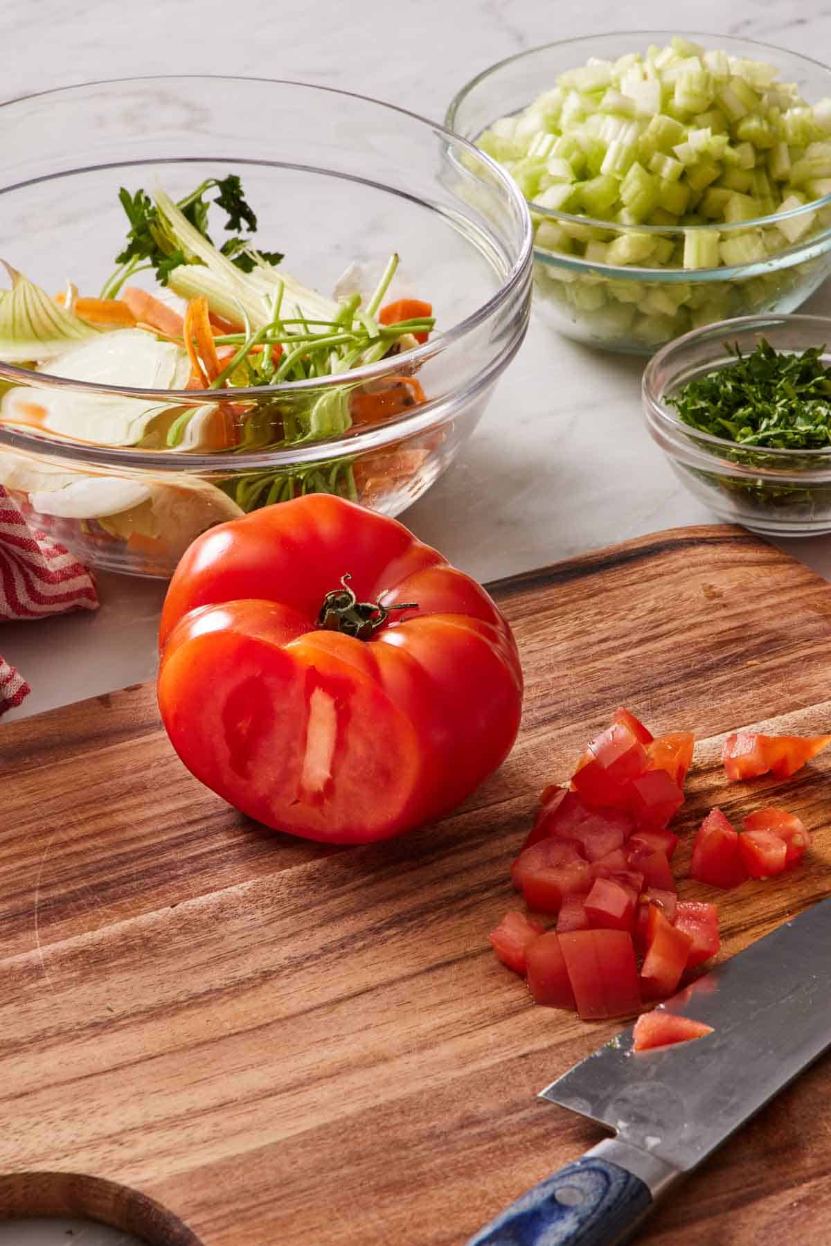 A partially chopped tomato on a wooden cutting board with a trash bowl and other chopped vegetables behind it.