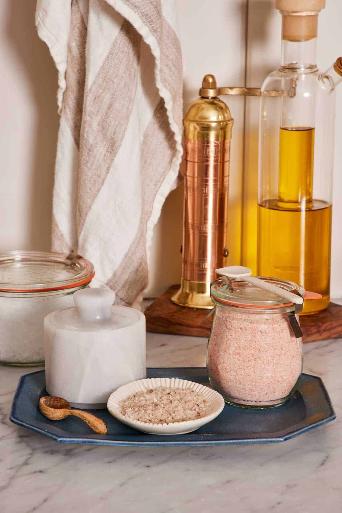 Fine and coarse pink salt in a jar and small bowl respectively on a blue platter with oil, a pepper mill, and sea salt in the background.