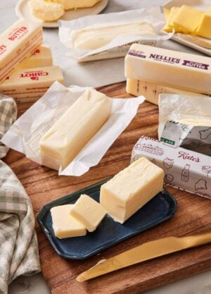 Various brands of butter sticks on a wooden cutting board and marble countertop, with one stick on a butter dish with pats cut off and stacked next to it.