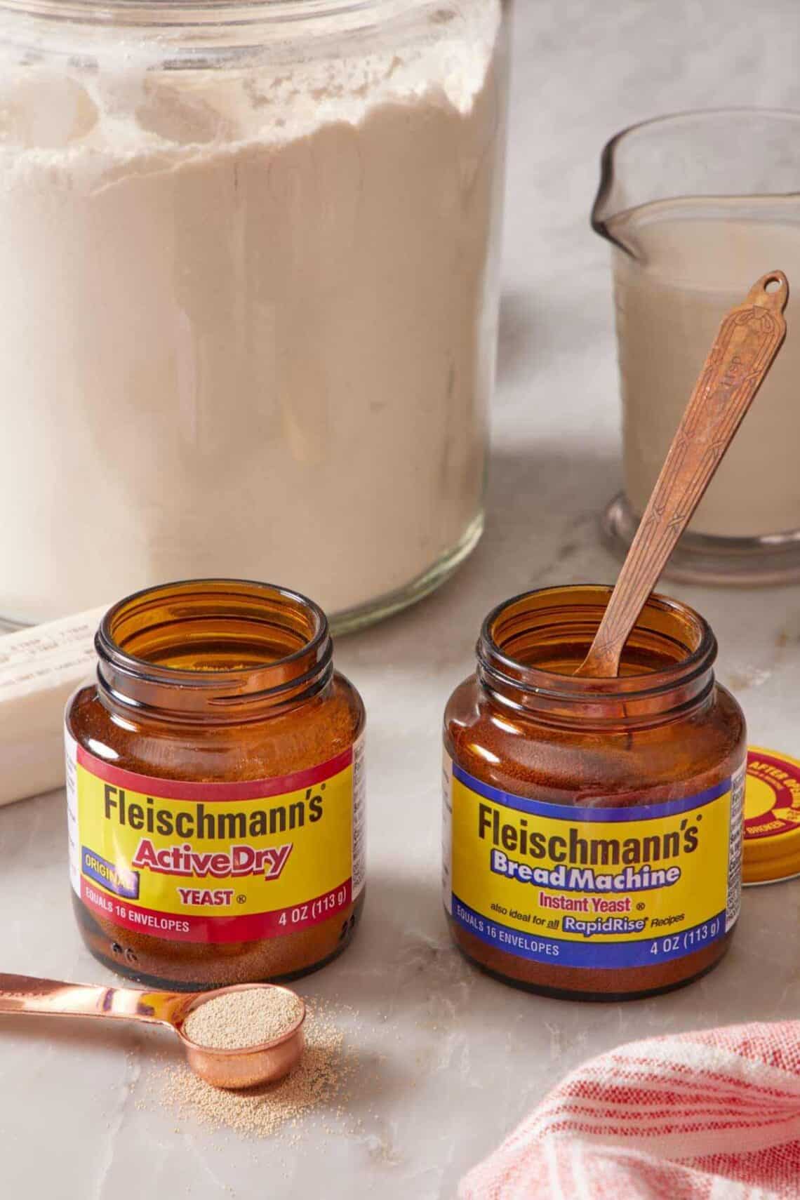 Two jars of different yeast side by side with a spoon in one jar and a spoon next to the other. Flour and a measuring cup of milk behind.