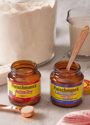 Two jars of different yeast side by side with a spoon in one jar and a spoon next to the other. Flour and a measuring cup of milk behind.