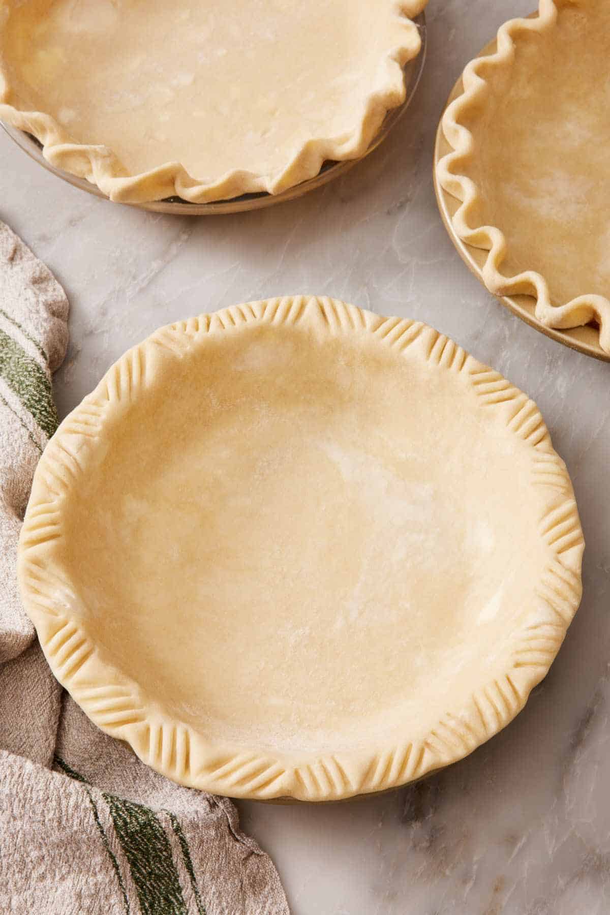 A pie crust in a pie dish with a fork crimped crust. A crust with a fluted edge and rope edge nearly out of frame.