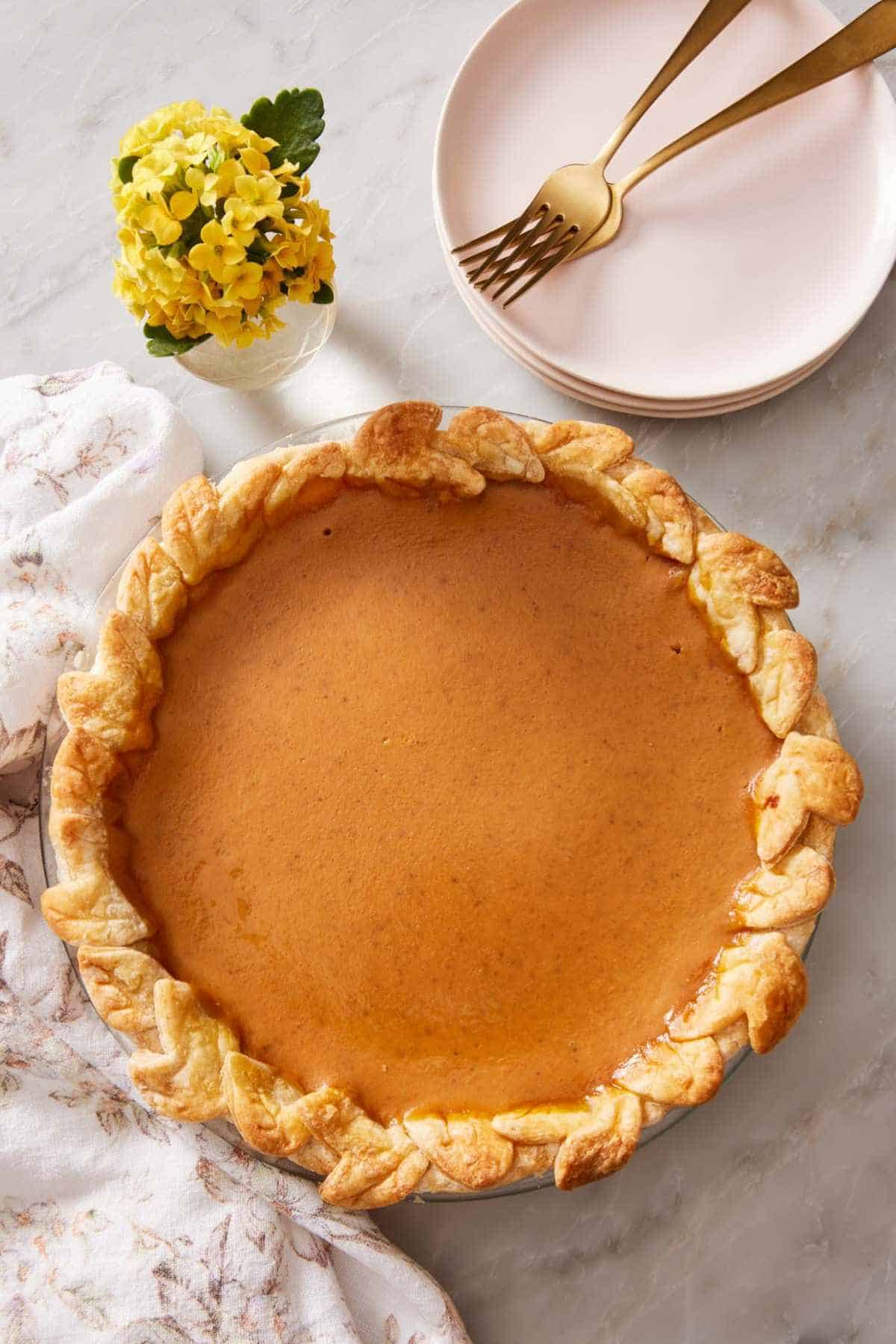 Overhead shot of a pumpkin pie with the edge decorated with cutouts. A few plates with forks on top off to the side.