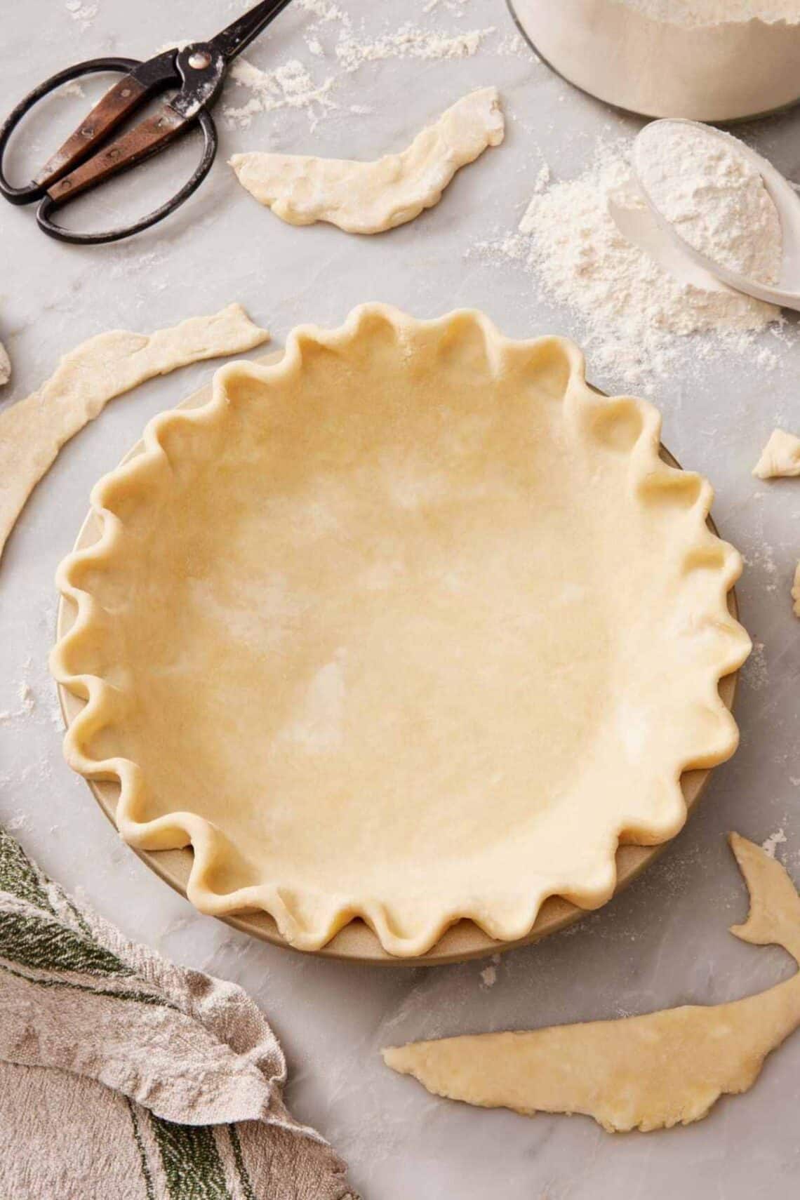 A pie crust in a pie dish with a classic crimp. Leftover pieces of pie dough scattered around.