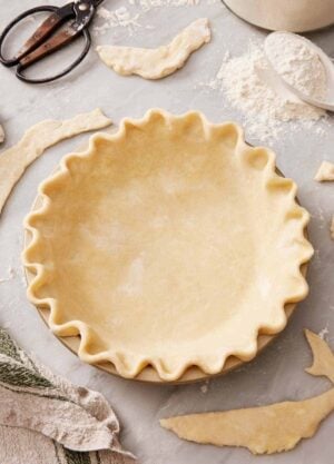 A pie crust in a pie dish with a classic crimp. Leftover pieces of pie dough scattered around.