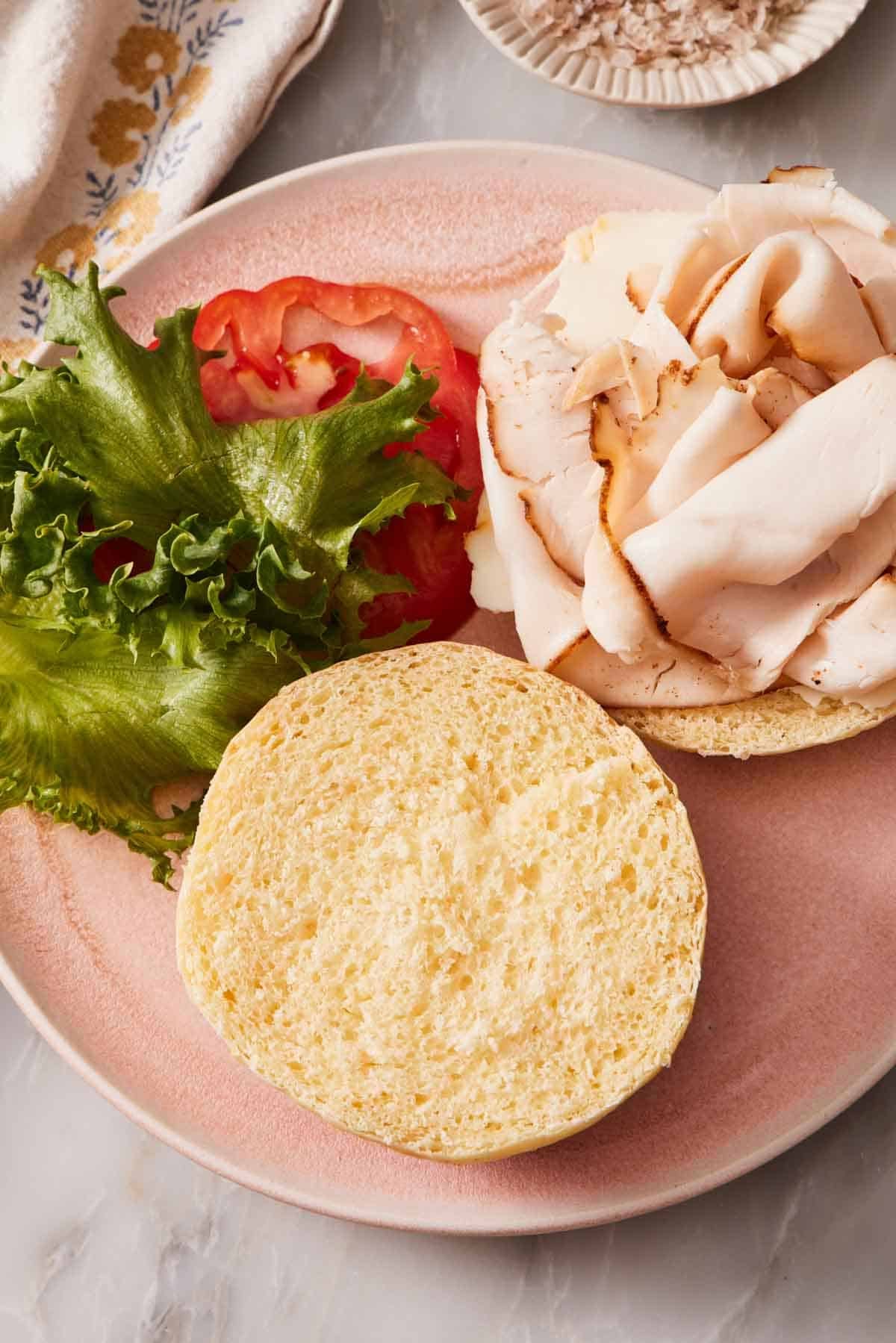 Overhead shot of a deli meat sandwich on a plate with the top of the bun taken off and placed next to it. Tomato and lettuce also on the plate.