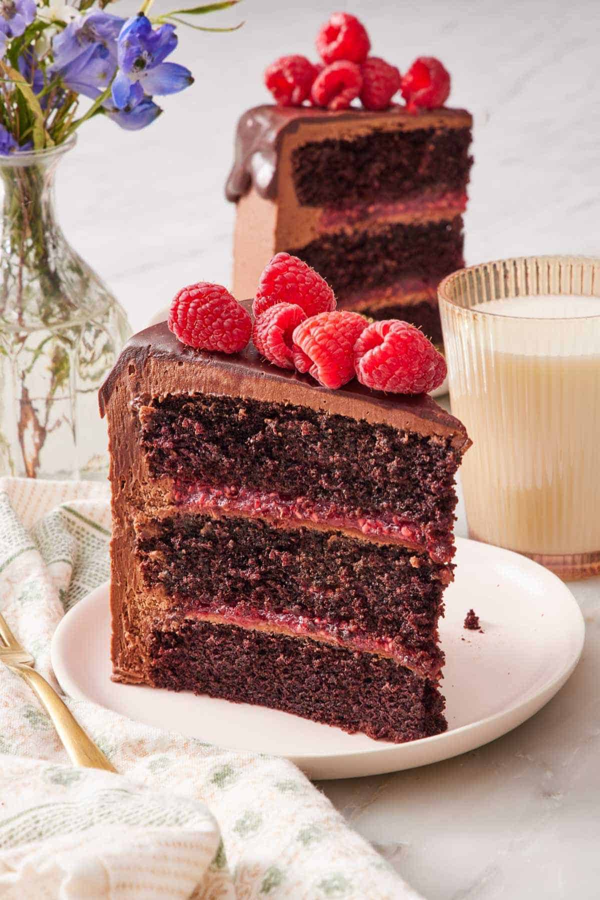 A slice of three-layer chocolate raspberry cake on a small plate with a glass of milk and another slice of cake behind it.