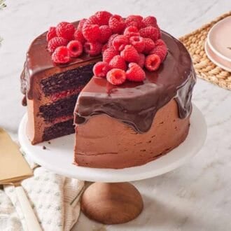 Chocolate raspberry cake on a cake stand with a cake server, a vase of flowers, and serving plates around it.