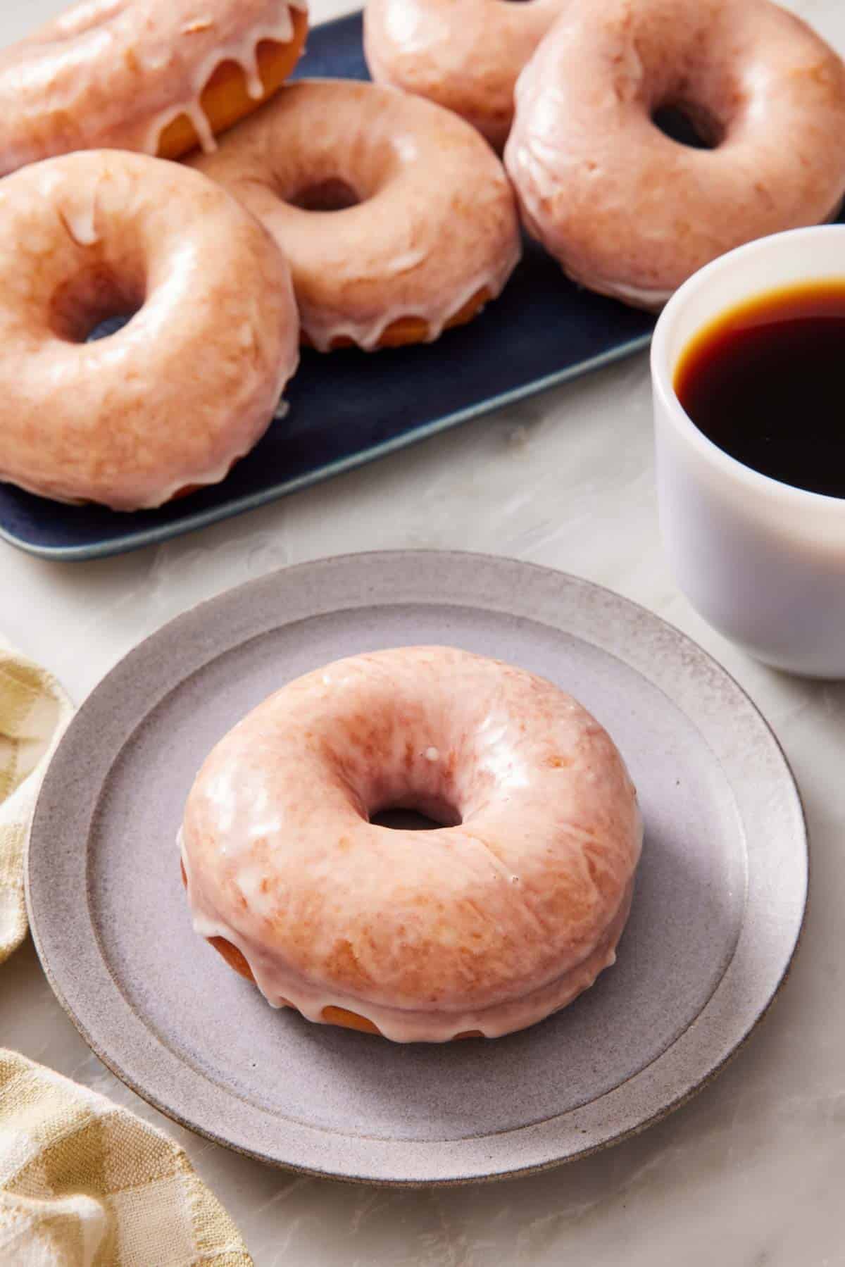 A glazed donut on a small plate alongside a cup of coffee, with more donuts in a platter behind.