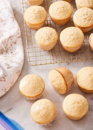 Vanilla cupcakes on a wire rack, as well as a few on a counter wrapped in plastic wrap.