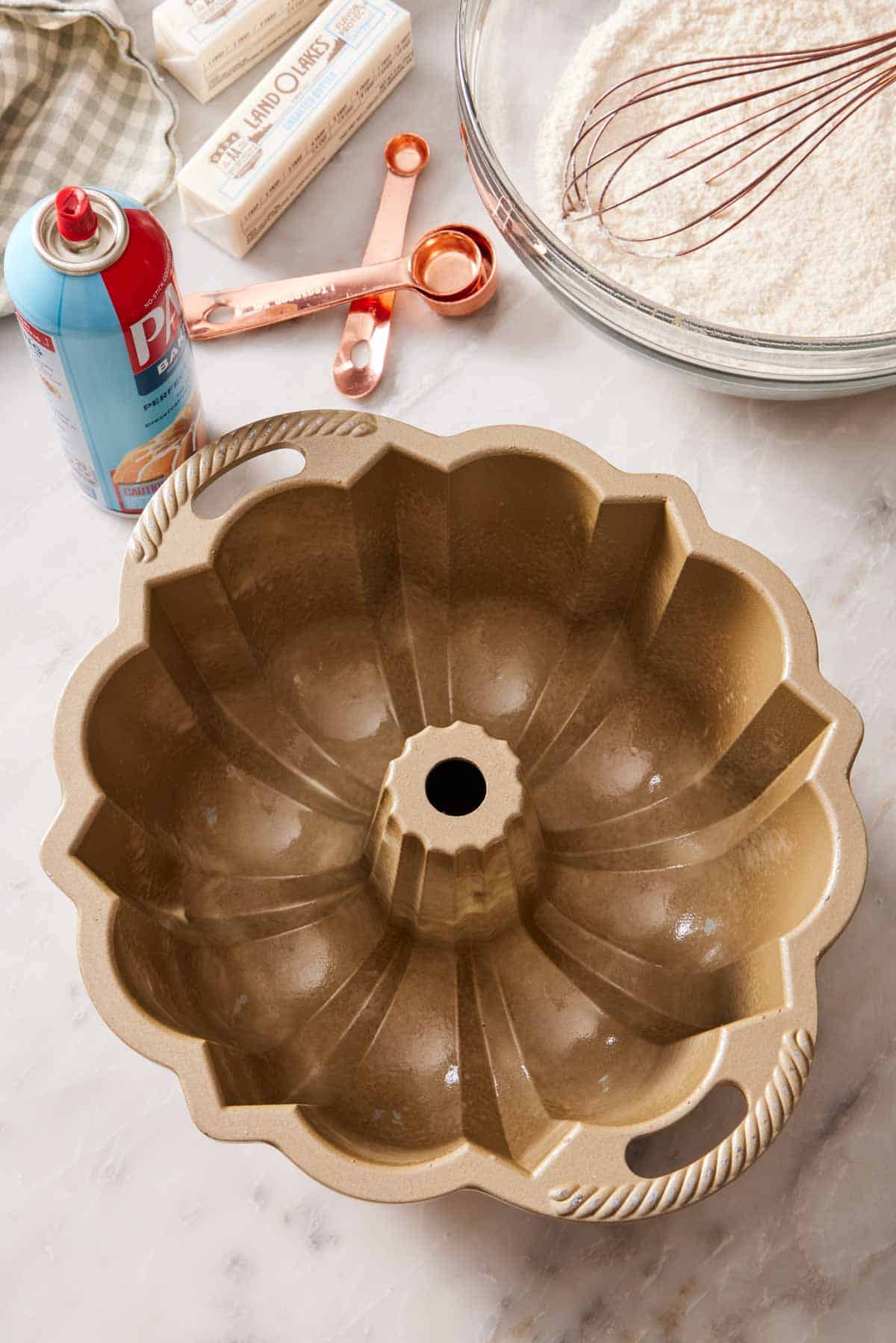 A greased Bundt pan with baking spray, measuring spoons, butter, and a bowl with flour behind it.