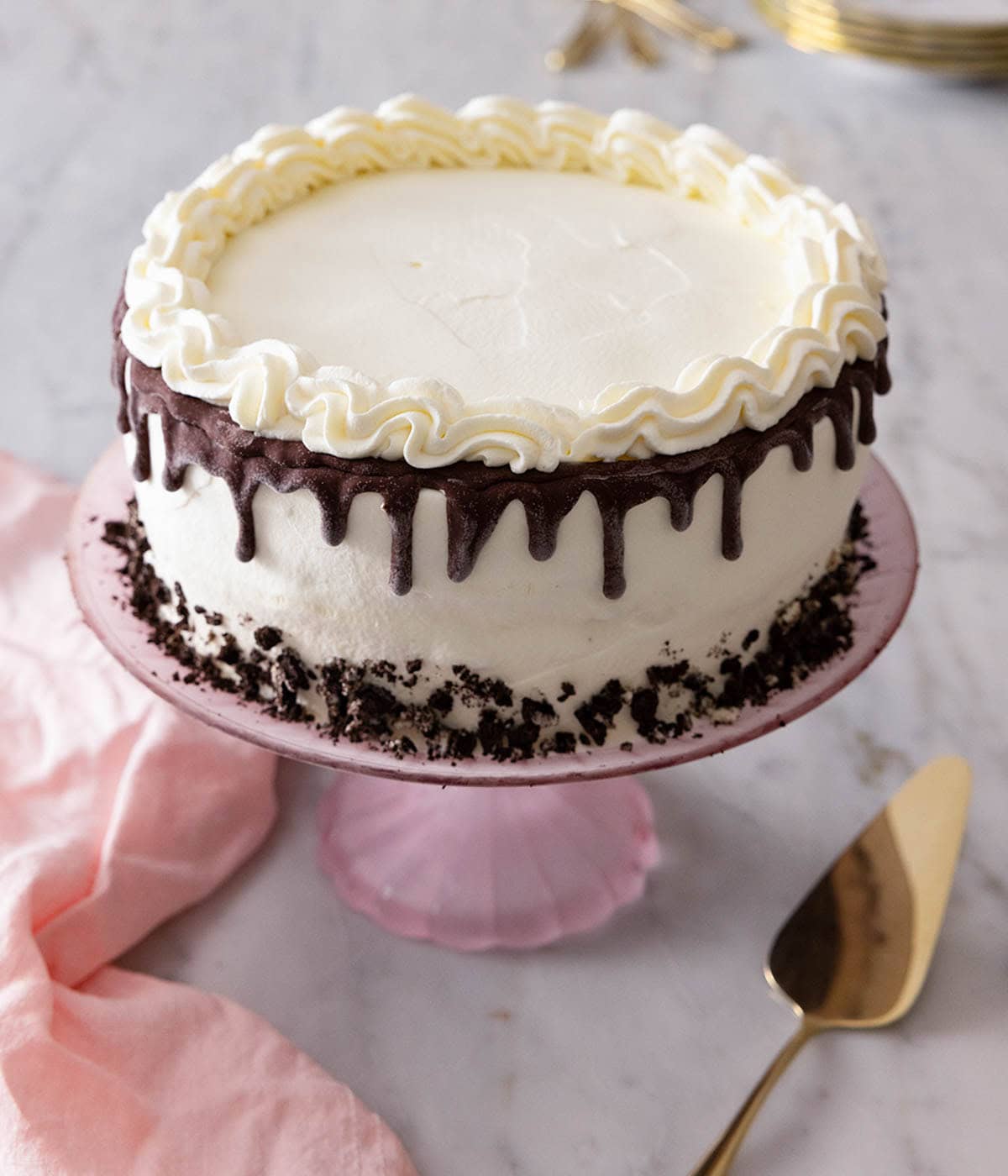 An ice cream cake on a cake stand with a cake server and pink table linen alongside.