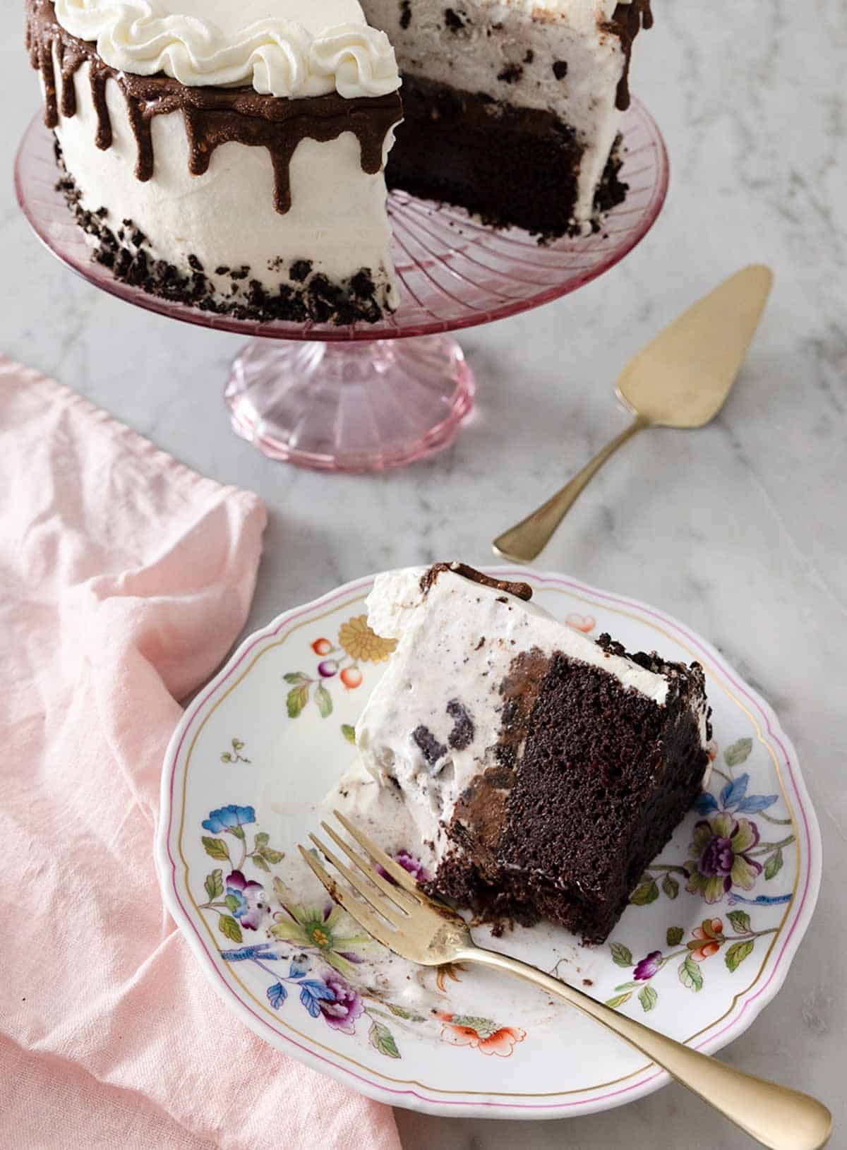 A slice of ice cream cake on a small plate with the rest of the cake on a cake stand behind it.