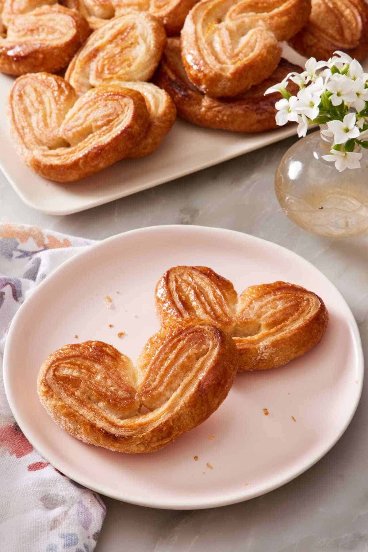 Close-up of two heart-shaped palmiers on a plate, with the rest on a platter behind them.