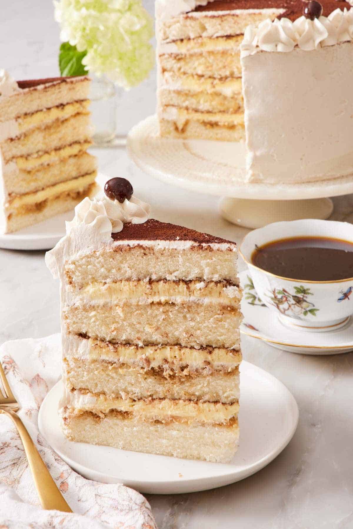 A slice of four-layer tiramisu cake on a plate with a cup of coffee, another slice, and the rest of the cake in the background.