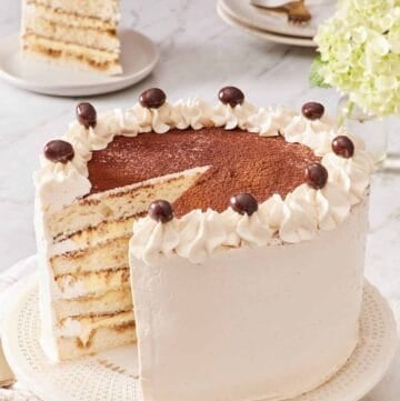 Tiramisu cake on a cake stand with a slice removed and placed on a plate in the background.