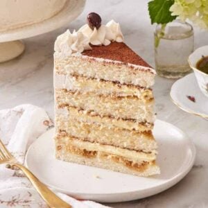 A slice of tiramisu cake on a small cake with a fork and a cup of coffee on either side.