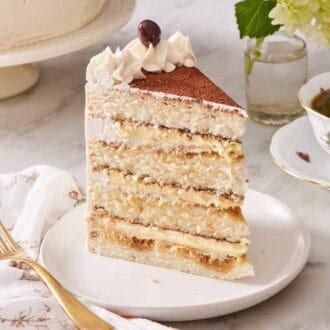 A slice of tiramisu cake on a small cake with a fork and a cup of coffee on either side.