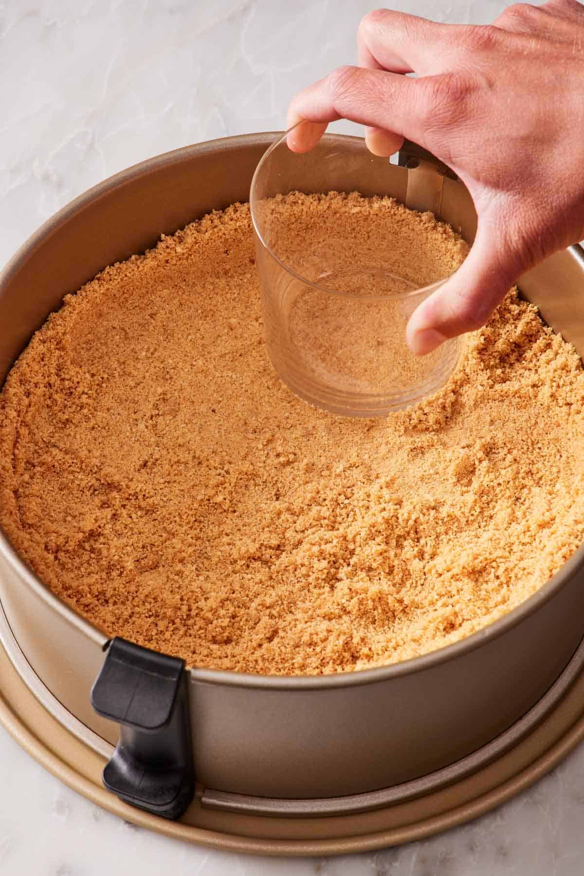 A graham cracker crust pressed into a springform pan with a flat-bottomed glass.