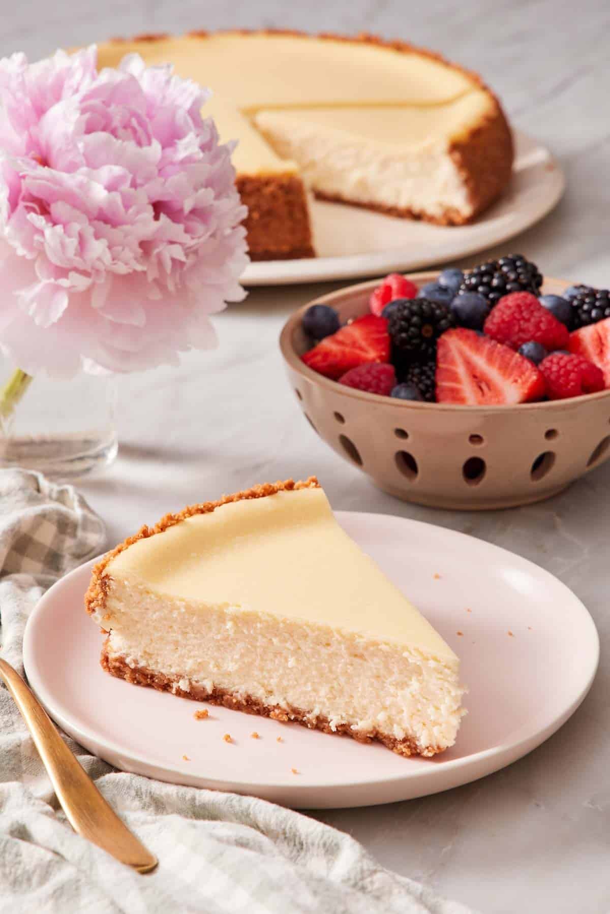 A slice of cheesecake on a plate with a bowl of berries and the rest of the cheesecake in the background.