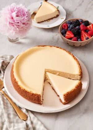 A cheesecake with one slice cut and placed on a plate, and the other slice still with the rest of the cheesecake. A bowl of berries and vase of flowers also in frame.