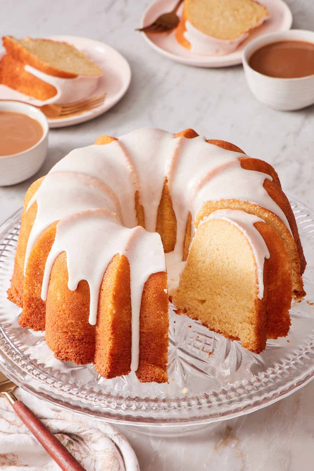 An iced Bundt cake on a cake stand with a few slices cut out and placed on plates in the background.
