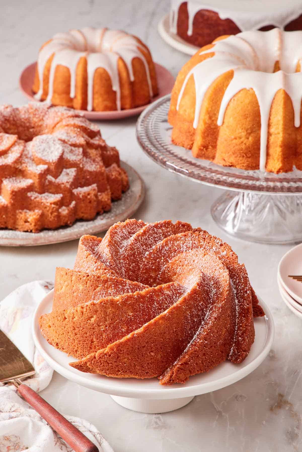 Different Bundt cakes on cake stands and plates set on a marble countertop.