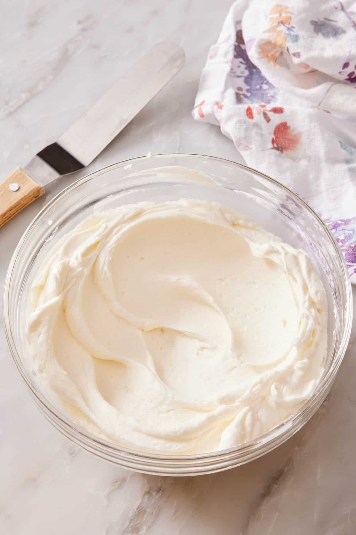 Buttercream frosting in a bowl on a marble countertop with an offset spatula and table linen alongside.