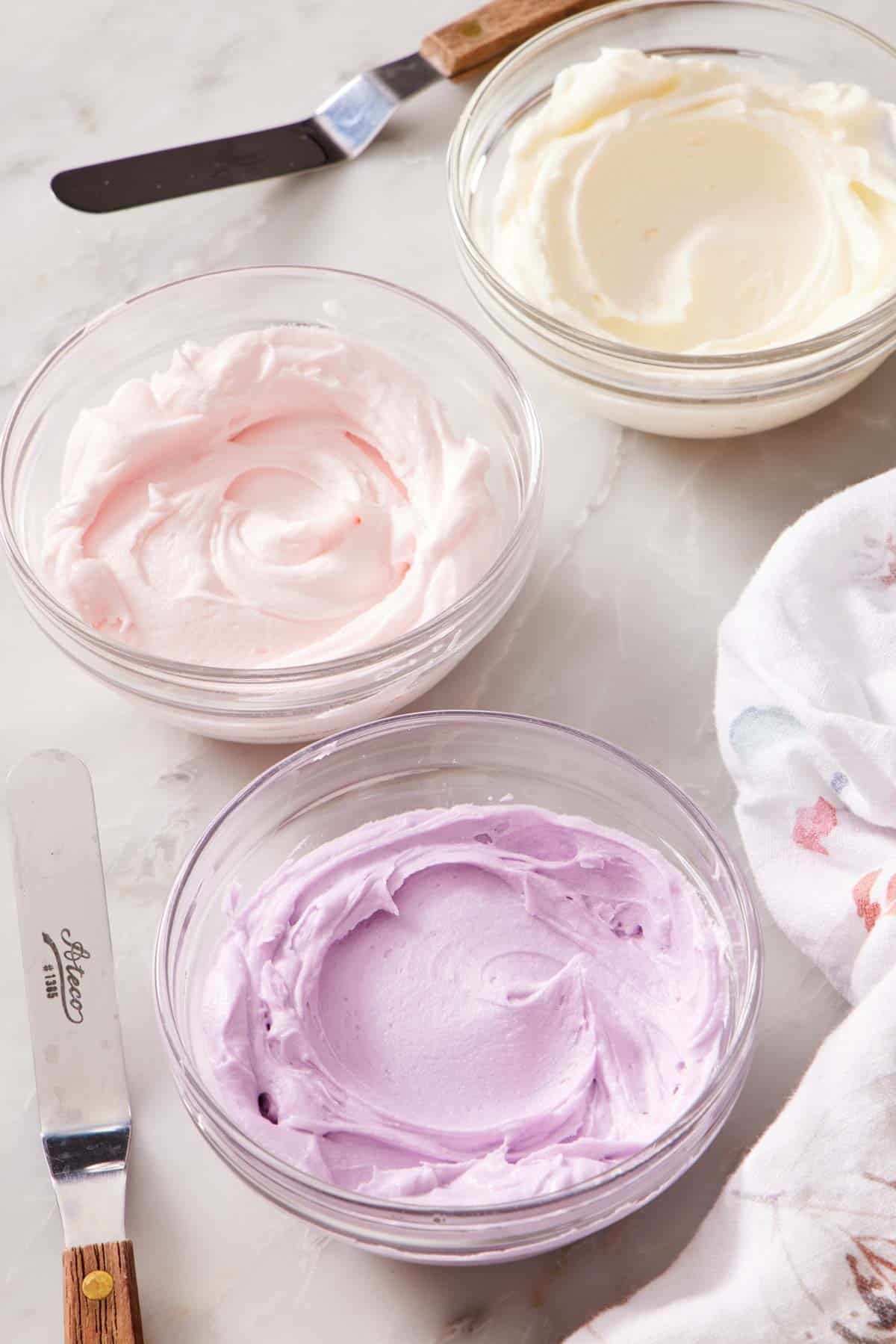 Buttercream frosting in three different colors in bowls on a marble countertop.
