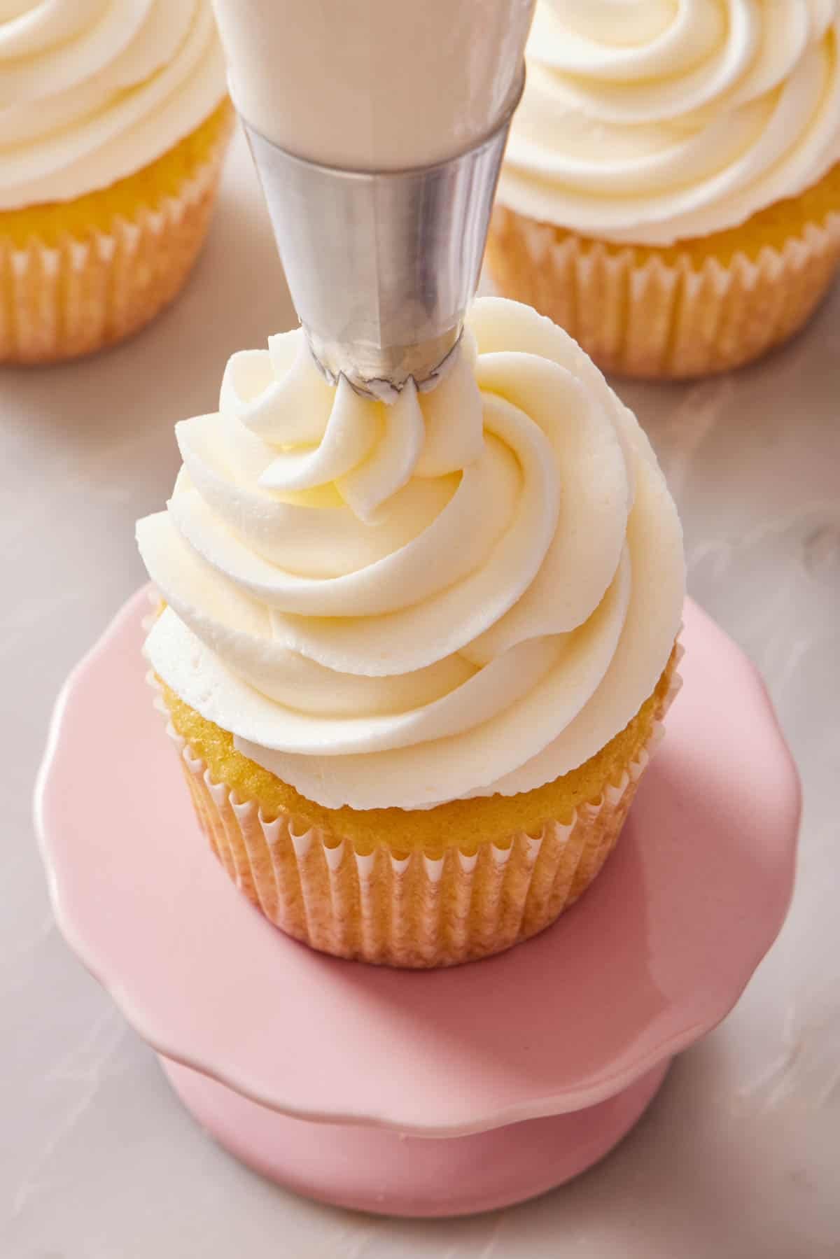 A cupcake on a small cake stand with frosting being piped on top.