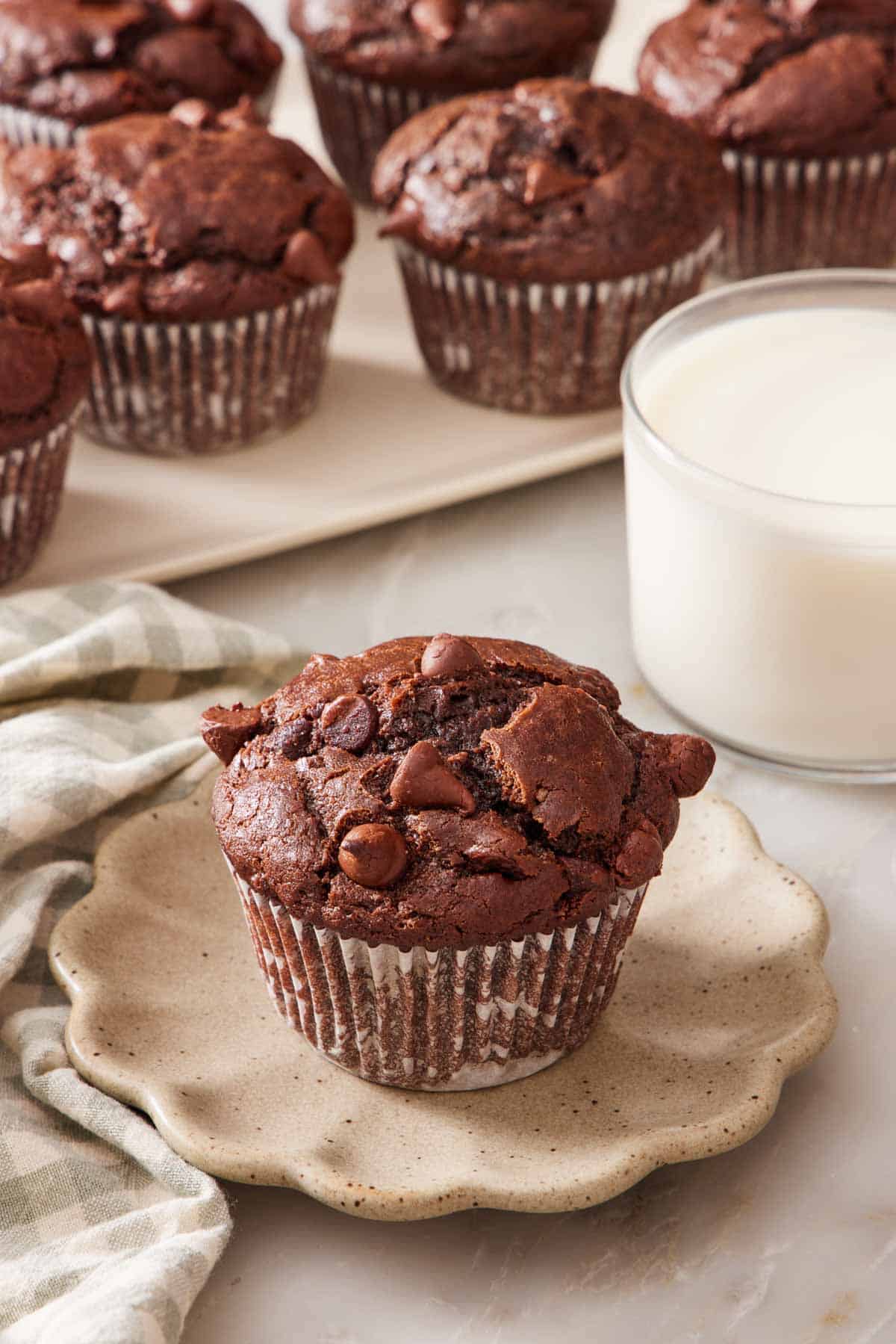 A chocolate muffin on a plate next to a glass of milk, with a platter of chocolate muffins in the background.
