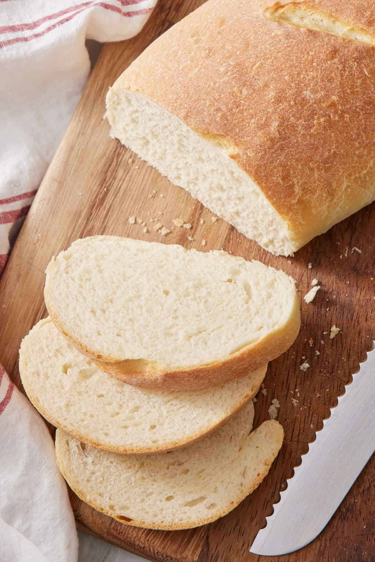 A loaf of Italian bread on a cutting board with a knife and three slices next to it.