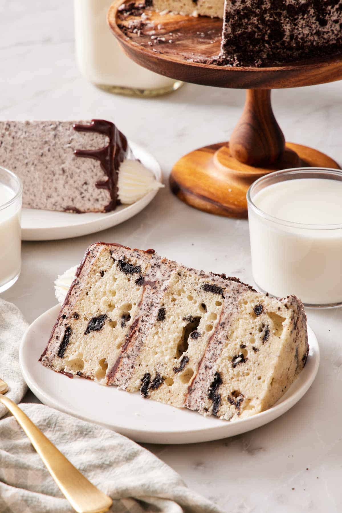 A slice of Oreo Cake on its side on a plate alongside a glass of milk. Another slice, glass, and the rest of the cake surround it but mostly out of frame.