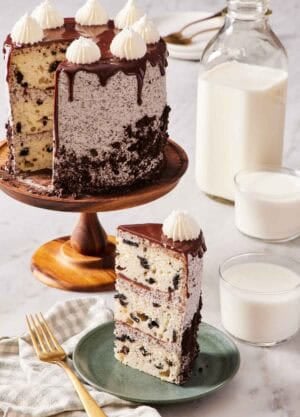 A slice of Oreo cake on a small green plate with the rest of the cake on a cake stand behind it. Two glasses of milk and also a jug of milk off to the side.