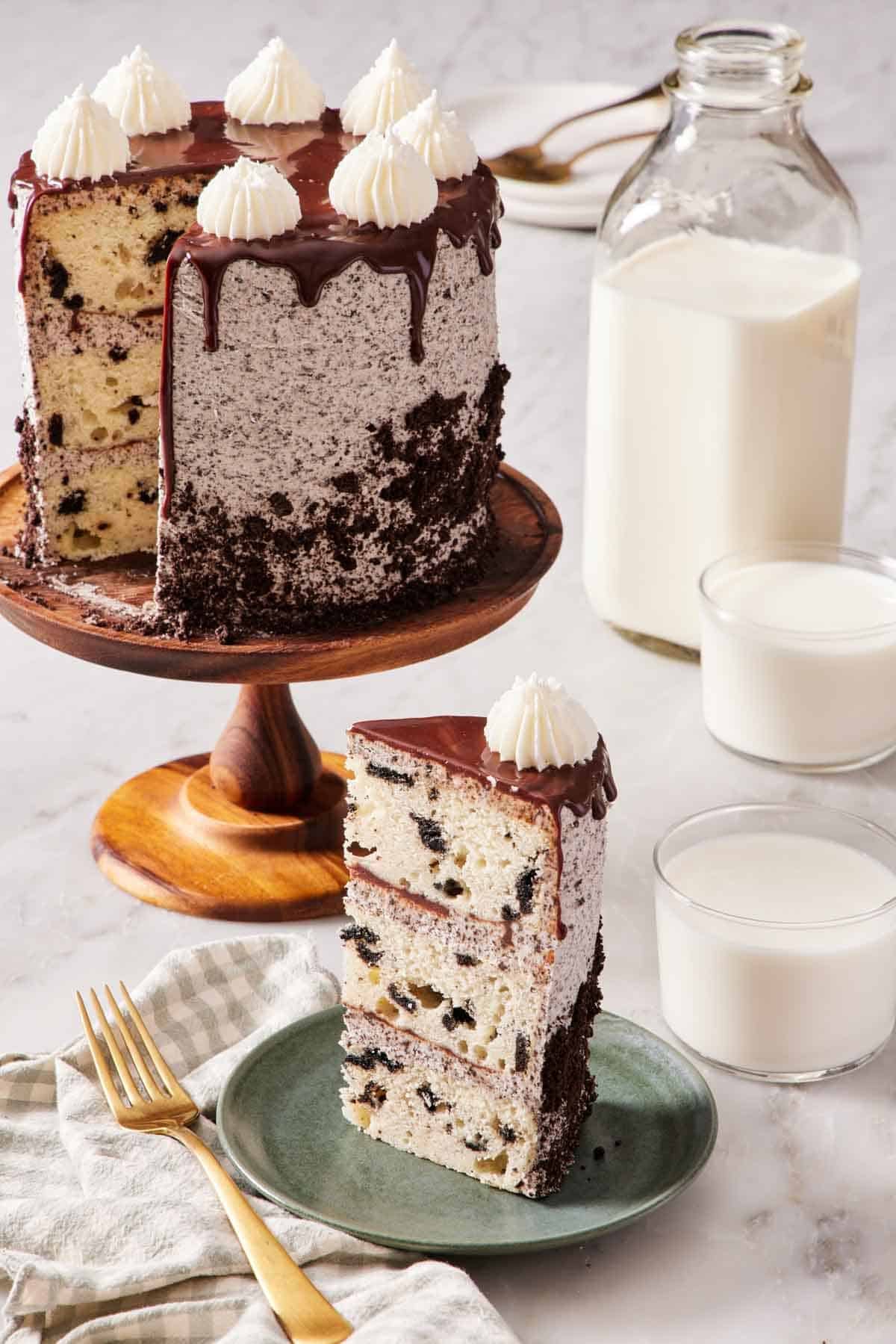 A slice of Oreo cake on a small green plate with the rest of the cake on a cake stand behind it. Two glasses of milk and also a jug of milk off to the side.
