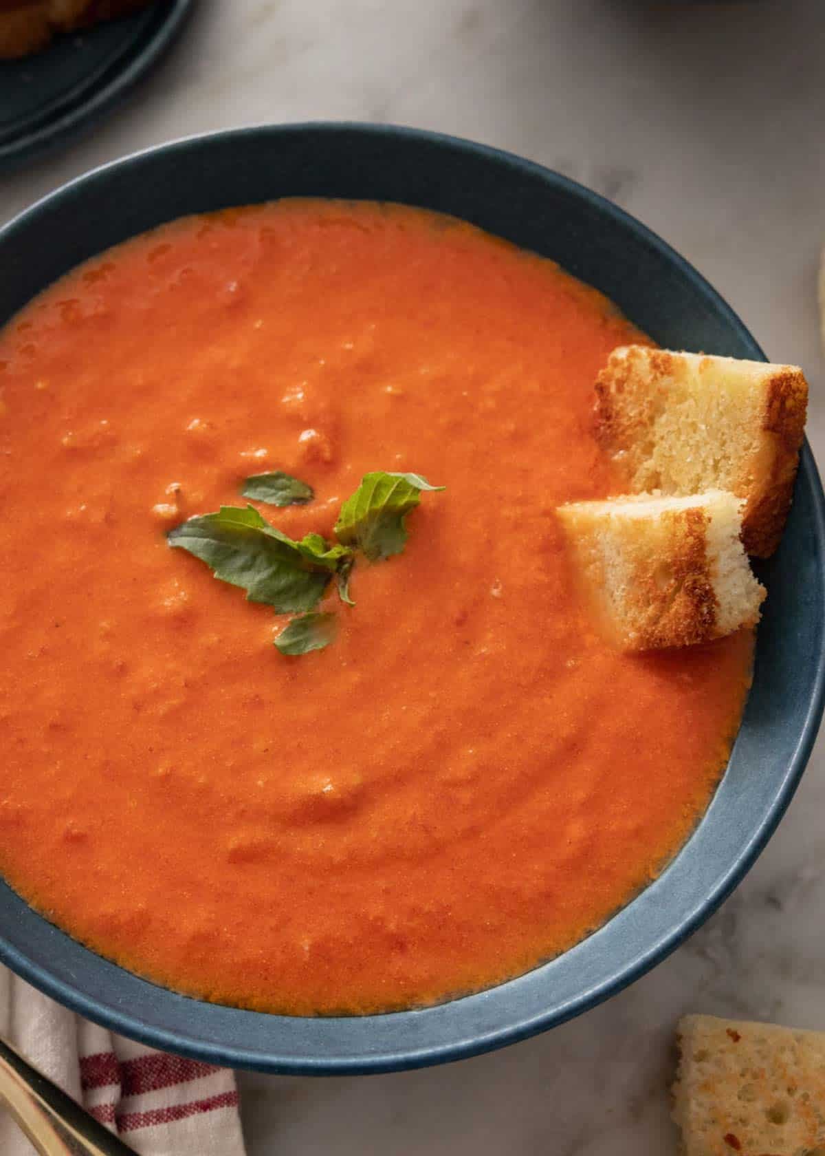 Overhead shot of tomato soup recipe in a bowl, with a few leaves of basil in the middle and toasted bread placed in the soup.