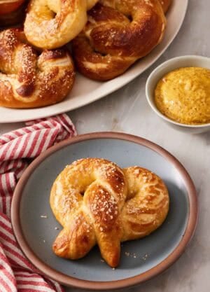 A soft pretzel on a plate with a dipping sauce and a pile of pretzels behind it.