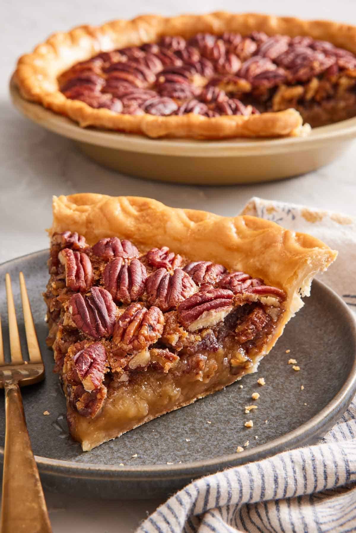 A slice of pecan pie in the foreground, with the rest of the pie out of focus in the background.