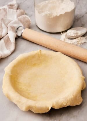 Pie dough laid in a pie dish with a rolling pin and a container of flour behind it.