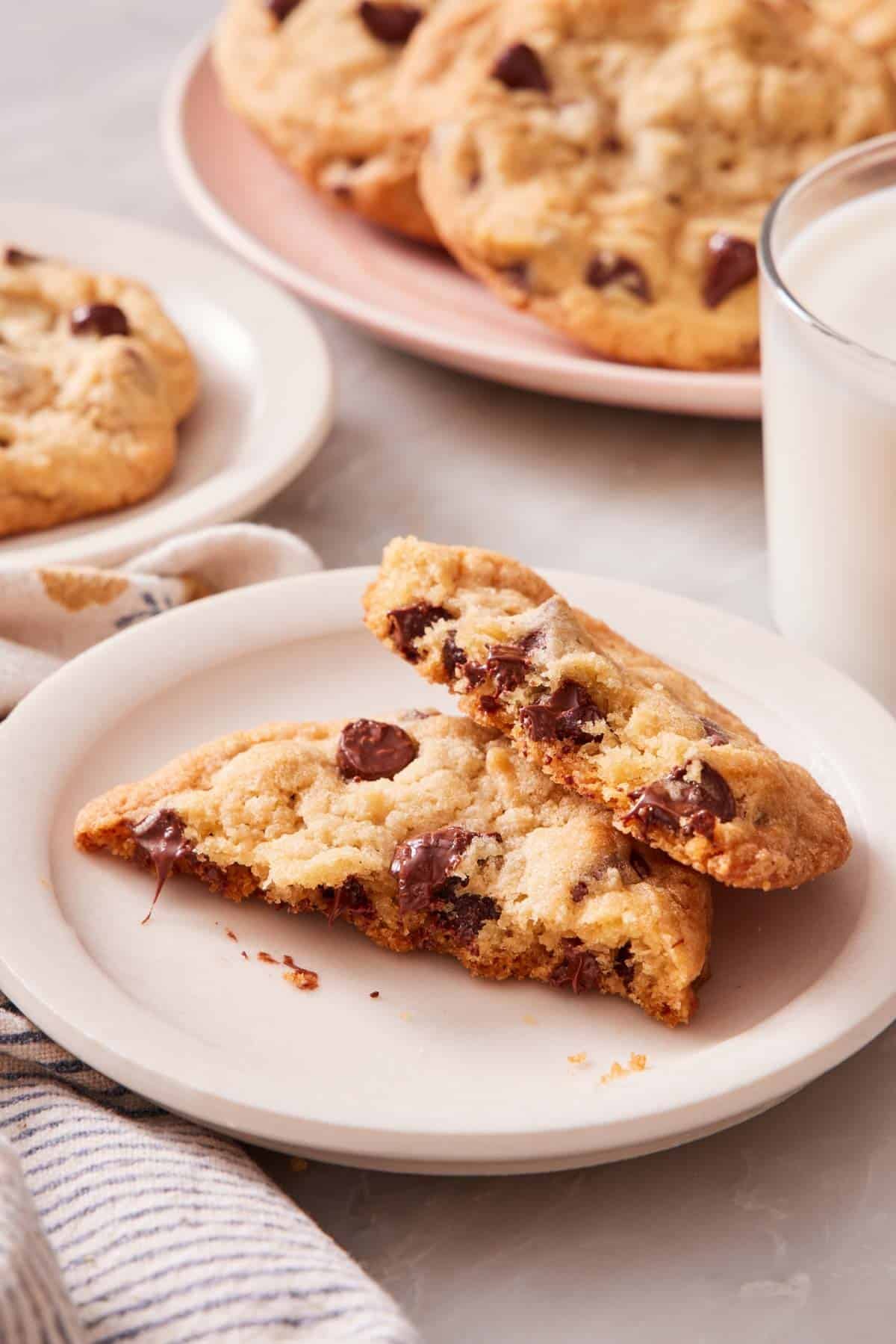 A cookie torn in half and placed on a small plate next to a glass of milk. More cookies in the background.