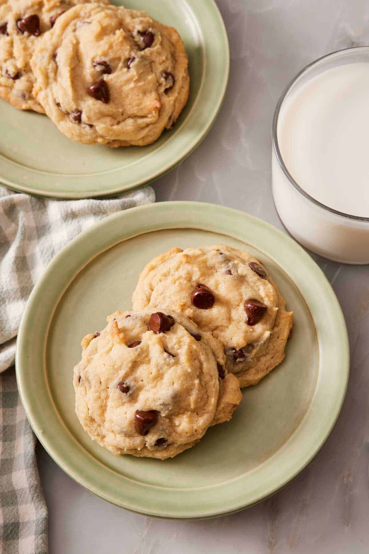 Overhead shot of two chocolate chip cookies on a green late next to a glass of milk. More cookies on another plate a little out of frame and out of focus as well.