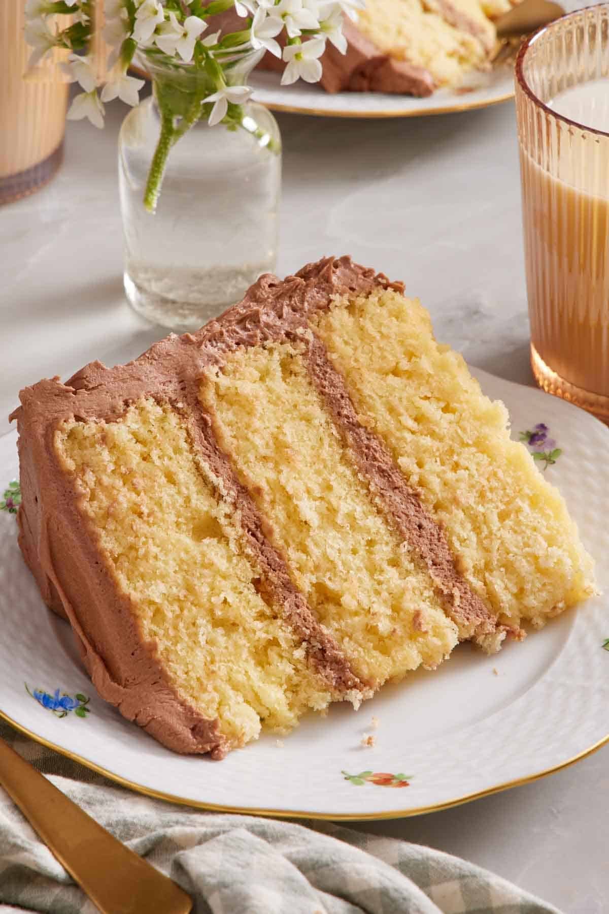 A slice of yellow cake on its side on a plate with a glass of milk next to it, mostly out of frame.