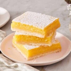 A stack of lemon bars on a small plate next to a table linen and vase of flowers.