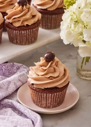 A mocha cupcake on a small plate with more cupcakes on a raised surface behind it, and a vase of flowers.
