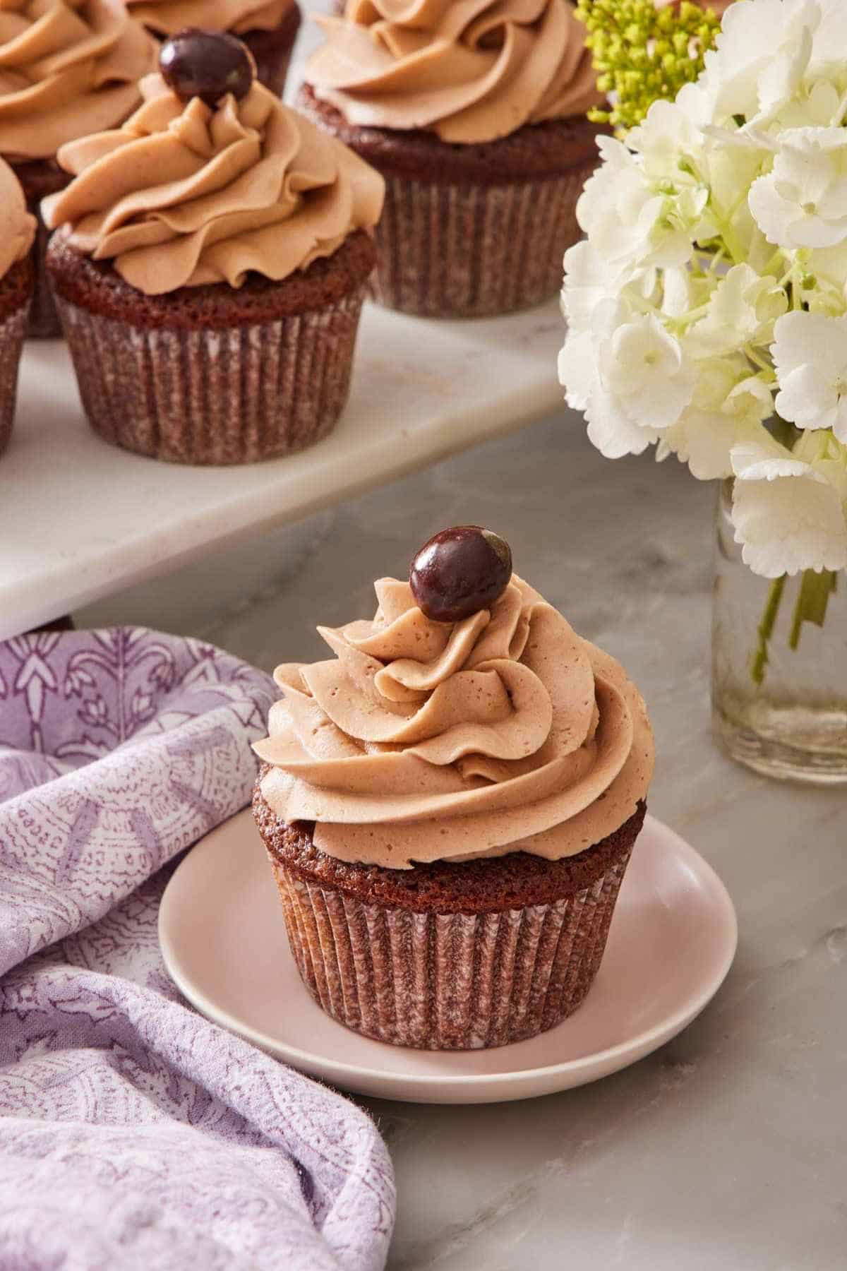 A mocha cupcake on a small plate with more cupcakes on a raised surface behind it, and a vase of flowers.