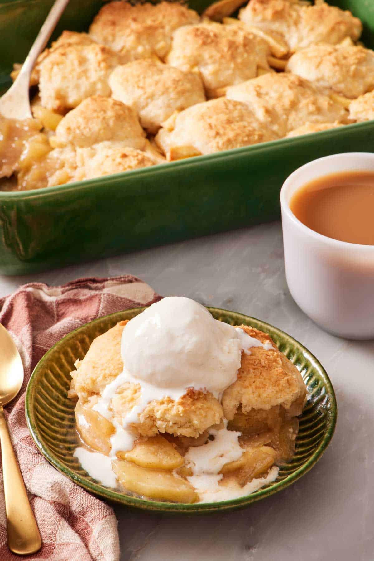 Apple cobbler in a bowl with ice cream with the rest of the cobbler in the background.