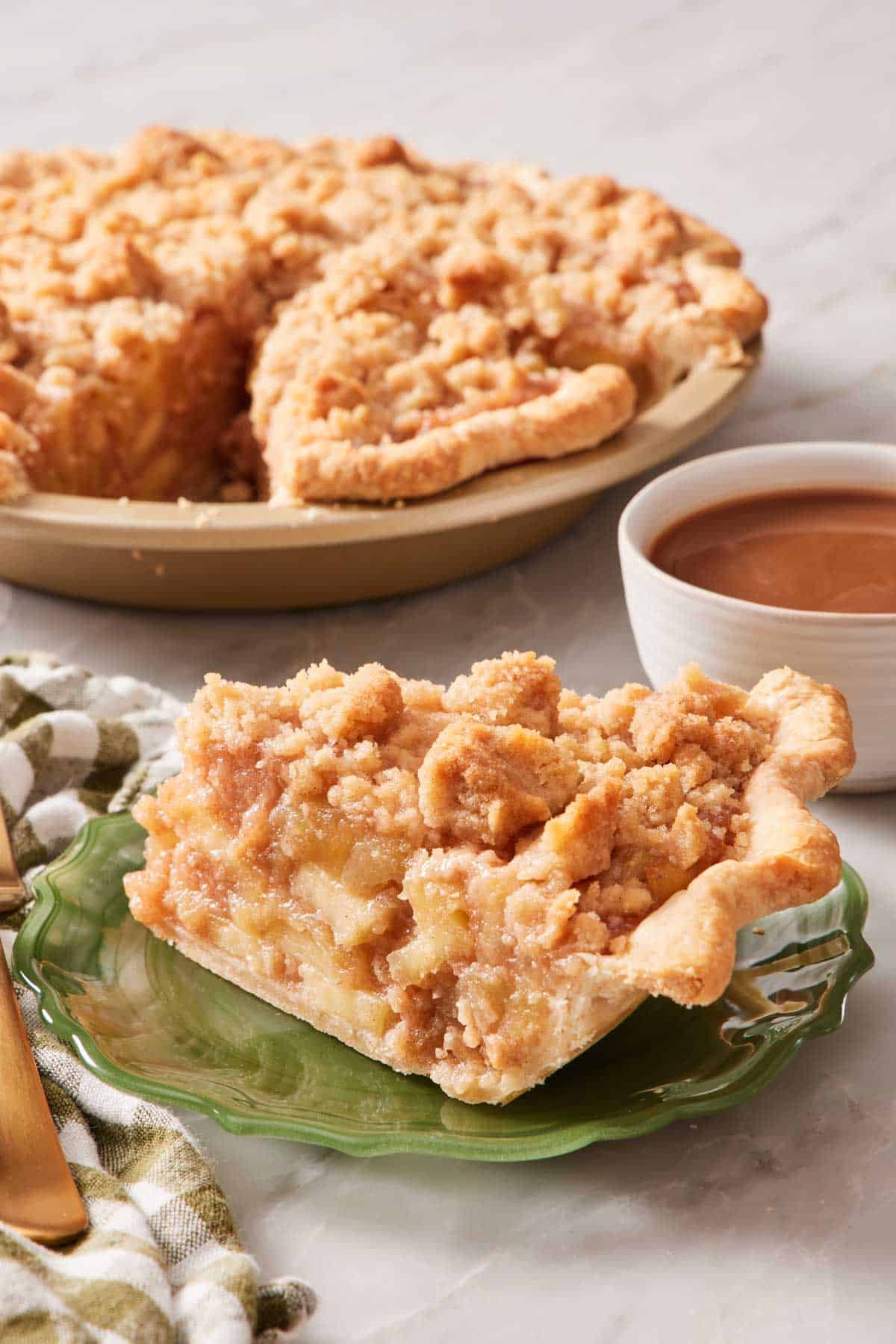 Slice of apple crumble pie (with homemade apple pie filling) on a plate with a drink and more pie in the background.