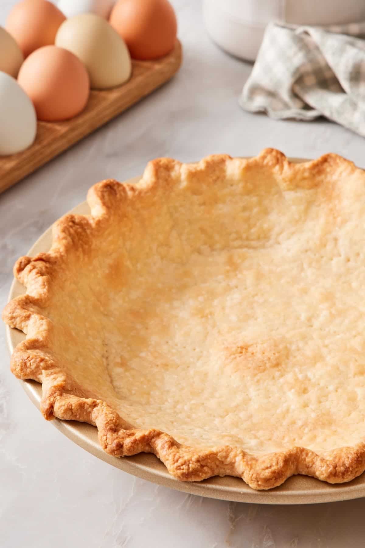Close-up of a blind-baked pie crust in a pie pan. Eggs in the background.